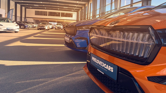 Rækker af biler holder parkeret i et solbeskinnet showroom med store vinduer og en nummerplade på en orange bil, hvor der står PRELOVEDCARS.