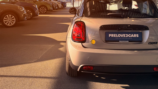 En sølvfarvet Mini Cooper med nummerpladen "Preloved Cars" holder parkeret blandt andre biler i solskin på en parkeringsplads.