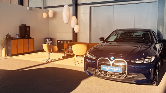 En marineblå BMW med PRE LOVED CARS-skilt er parkeret indendørs ved siden af et stilfuldt venteområde med stole, et bord og moderne hængelamper.