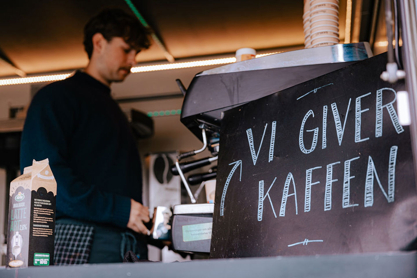 En person står bag en kaffemaskine ved siden af et skilt på dansk, hvor der står: »Vi giver kaffen«.