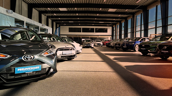 PreLovedCars showroom med rækker af forskellige brugte biler udstillet indendørs under naturligt lys fra store vinduer.