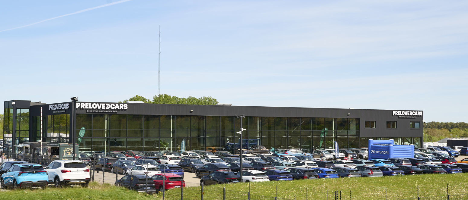 En stor bilforhandler med mange parkerede biler udenfor og en bygning med påskriften PRELOVEDCARS under en klar blå himmel.