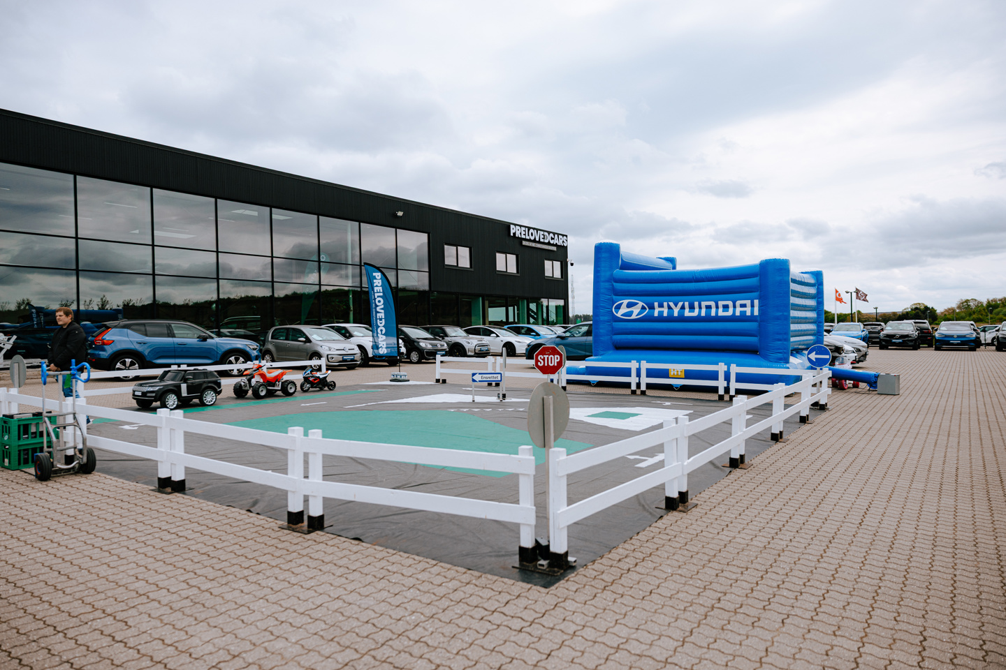 En Hyundai-forhandlers udstillingsplads med biler, en blå hoppeborg med Hyundai-logo og en indhegnet legeplads med legetøjsbiler og vejskilte.