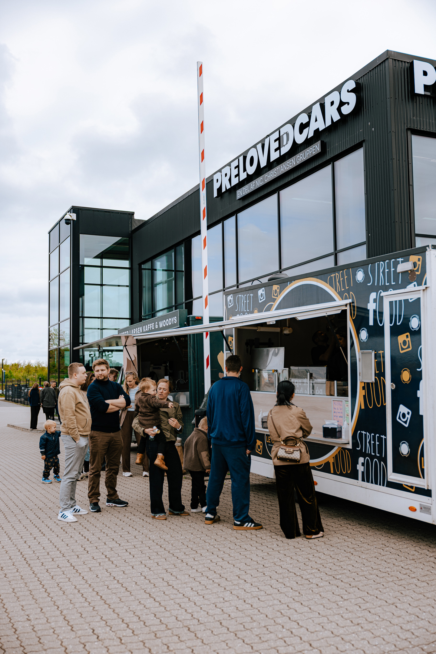 Folk står i kø ved en madvogn, der holder parkeret uden for en moderne bygning med skiltet PRELOVEDCARS.