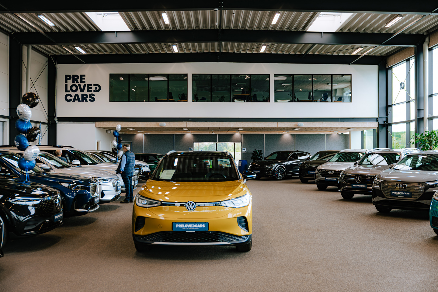 En gul Volkswagen står udstillet i et udstillingslokale sammen med flere andre biler, og på væggen hænger der et skilt med teksten »PRE LOVED CARS«.