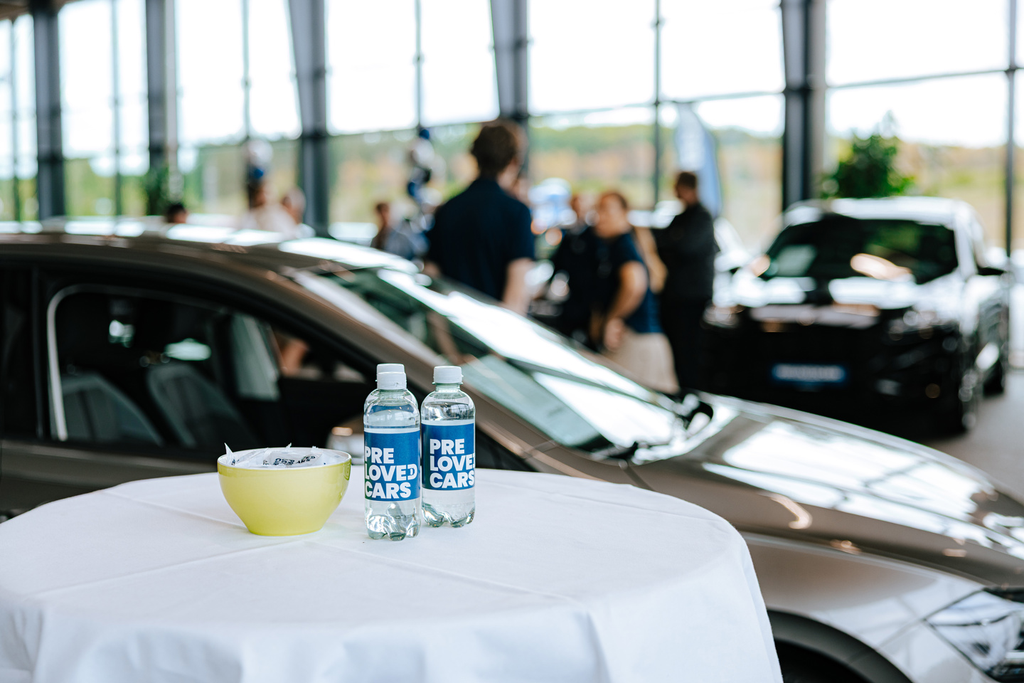 Et hvidt bord med to vandflasker med påskriften »Pre Loved Cars« og en skål står opstillet foran biler og mennesker inde i et udstillingslokale.