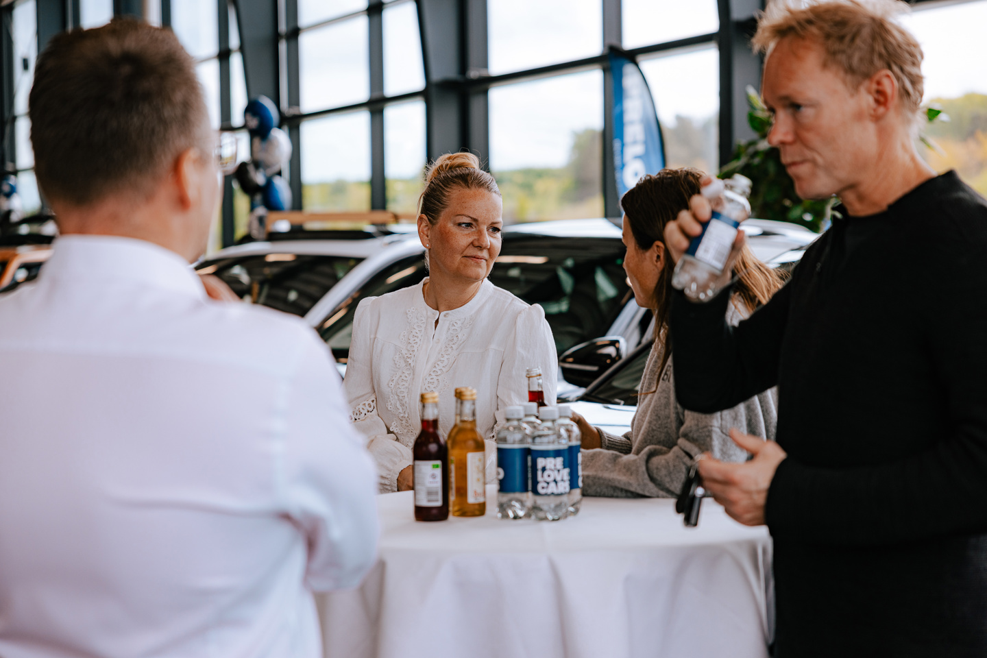 Fire personer står omkring et højt bord med drikkevarer på flaske og en krukke med pastiller i et biludstillingslokale.