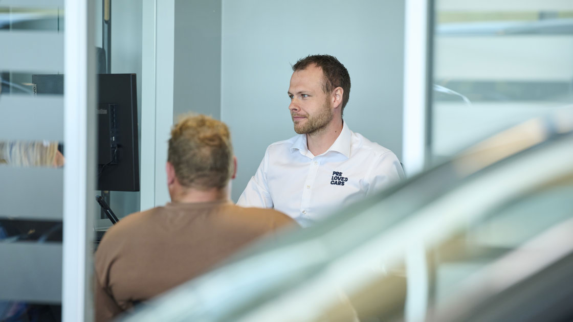 To mænd sidder over for hinanden ved et skrivebord på et kontor, den ene foran en computerskærm og den anden iført en hvid skjorte.