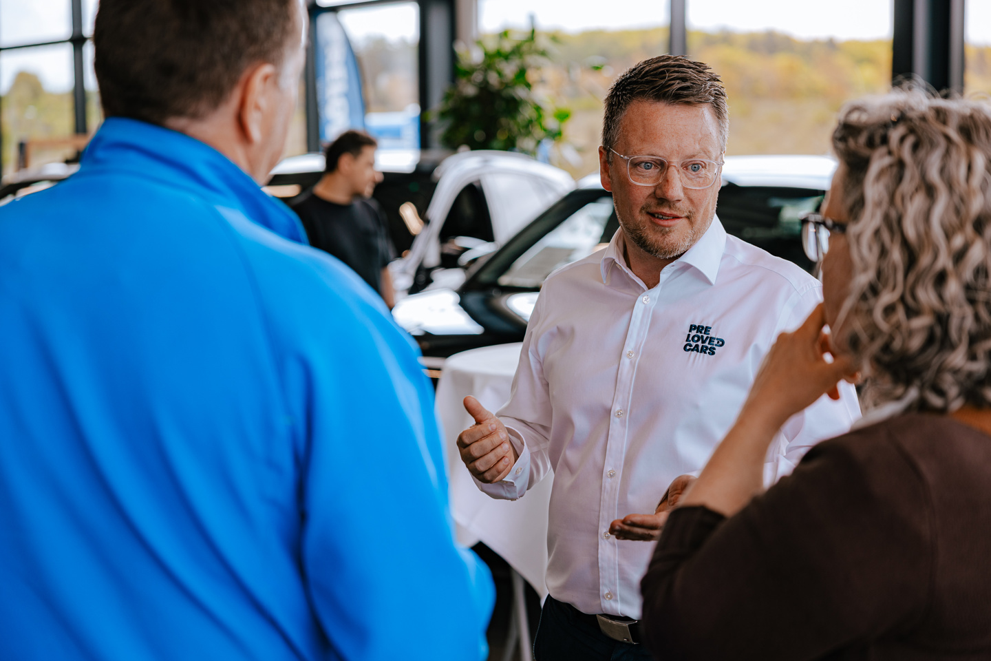 En mand i en hvid PRE LOVED CARS-trøje taler med to personer inde i et bilhus, hvor man kan se biler i baggrunden.